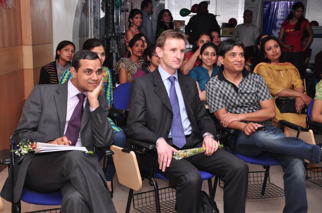 Swissnex delegate seated with Su-Kam hosts and staff at an indoor welcome event — flowers, blue lecture chairs, Gurugram