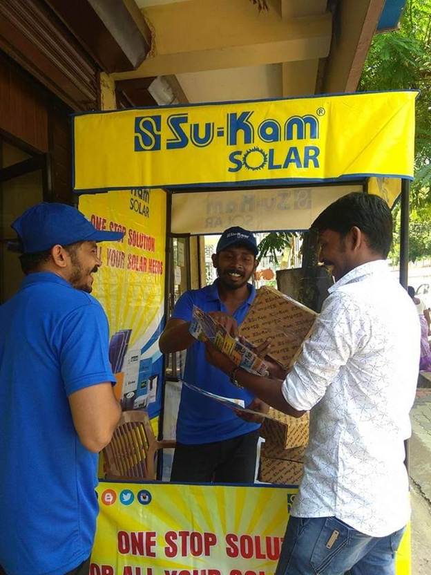Su-Kam staff handing product brochures to a customer at a Su-Kam Solar branded outlet with the company board overhead